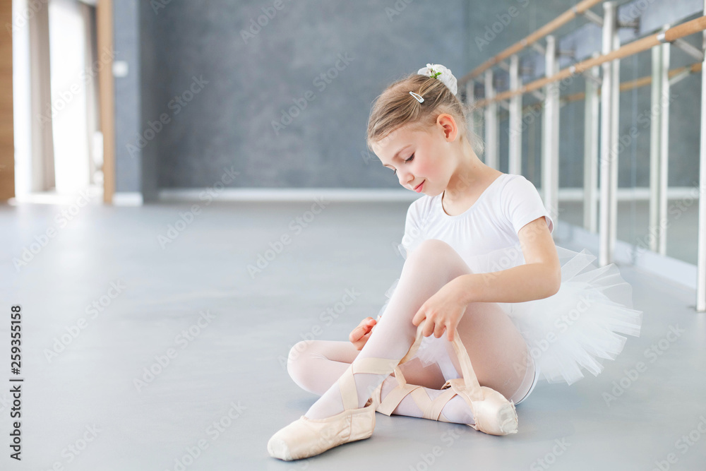 Little ballerina is trying on pointe shoes in ballet class. Cute kid girl  is sitting on