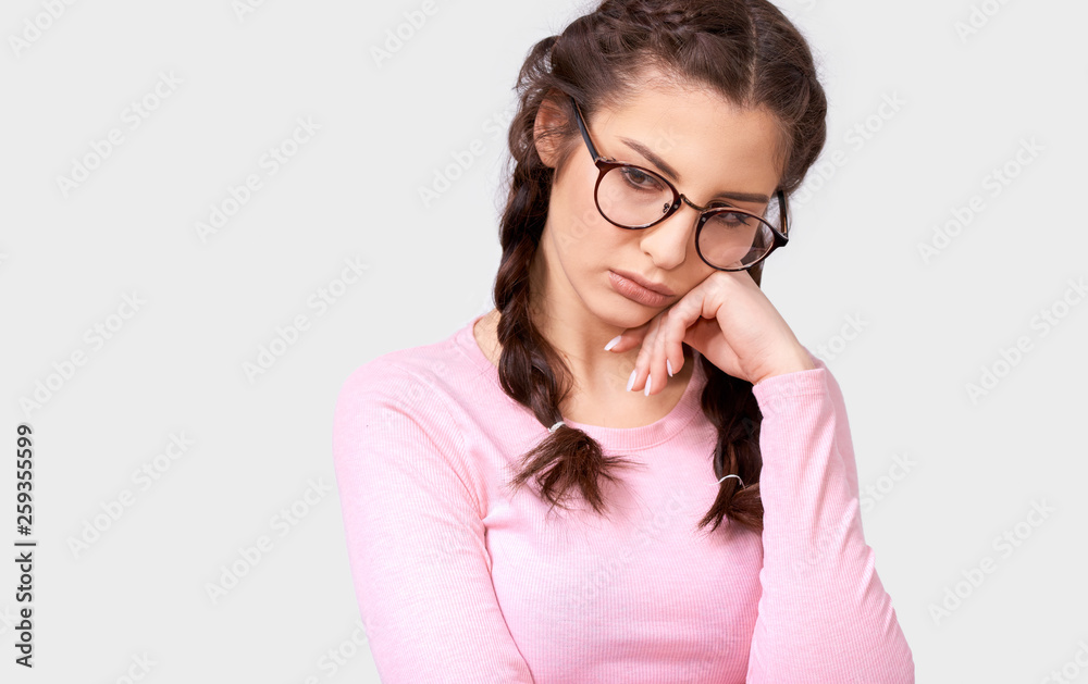 Close up portrait of overworked young brunette woman leans on hand, has tired facial expression, wears eyewear and pink casual blouse, isolated over white background. Shot of pretty sad female on gray