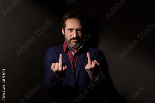 Model tests for men. A man in a suit cornered in black. Wearing a red shirt, blue jacket and showing middle fingers with both hands with aggression on his face