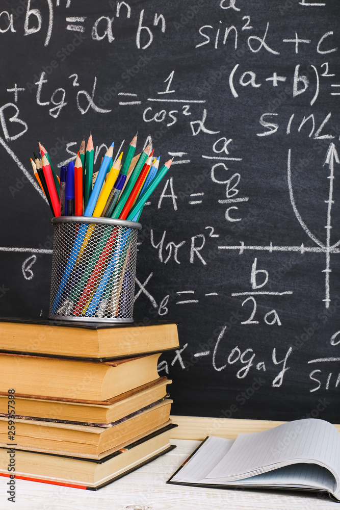 School desk in classroom, with books on background of chalk board with written formulas. Soncept Teacher's Day. School desk in classroom, with books on background of chalk board with written formulas. Soncept Teacher's Day.