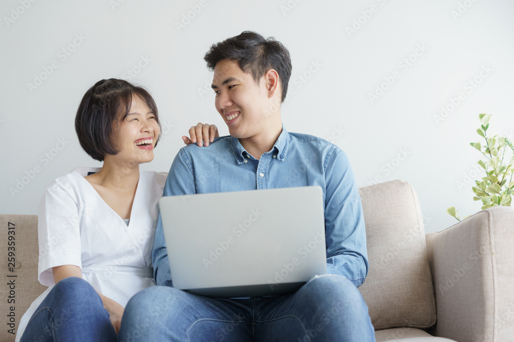 Naklejka premium Asian love couple sitting on the sofa and using laptop they is smiling happily at home