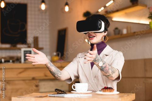 smiling businesswoman in virtual reality headset sitting at table with coffee cup and cake in cafe