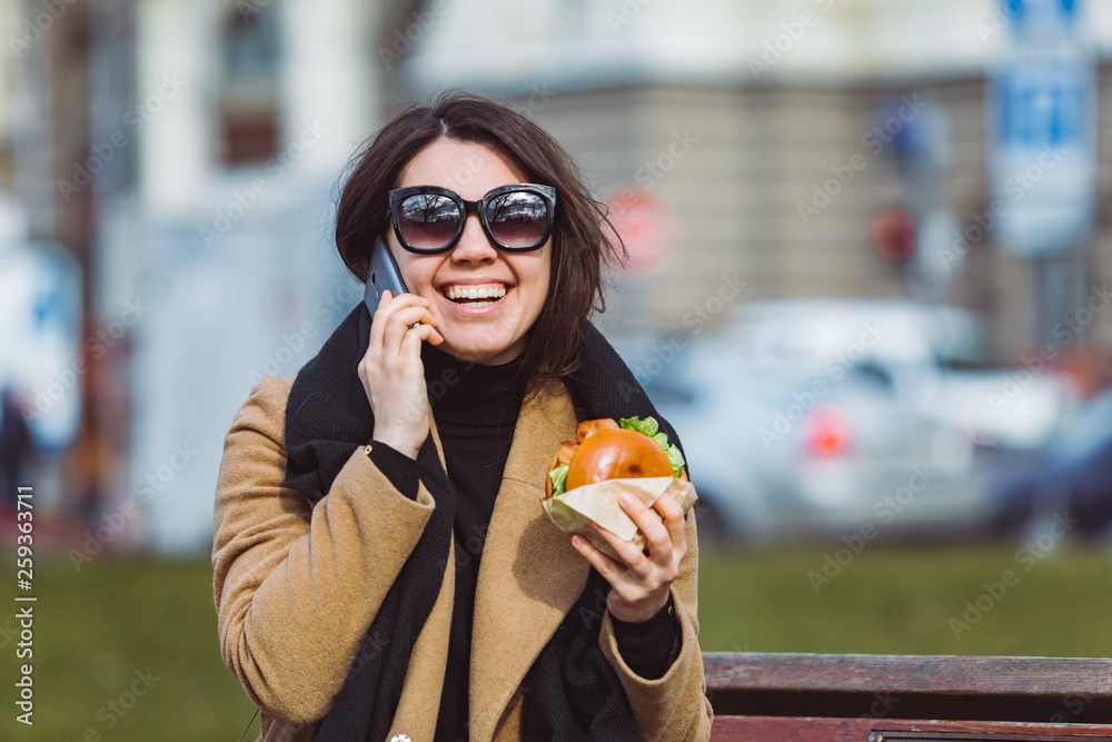 young beauty business woman eating fast food and working on phone while sitting at city bench