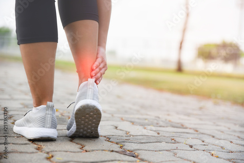 Woman hands holding her ankle while running on road in the park with red color highlight at ankle. Injury from workout concept.