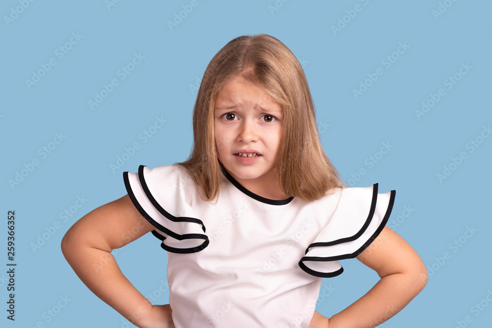 Close up emotional portrait of young blonde girl on blue background in studio. She holds her hands at the waist and is complaining about something or expresses dissatisfaction with something. Close up emotional portrait of young blonde girl on blue background in studio. She holds her hands at the waist and is complaining about something or expresses dissatisfaction with something.