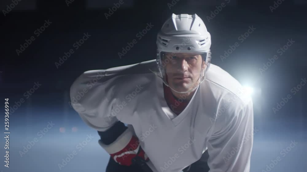 Portrait of determined middle-aged Caucasian ice hockey player in helmet, jersey and gloves standing on rink and looking at camera, medium shot