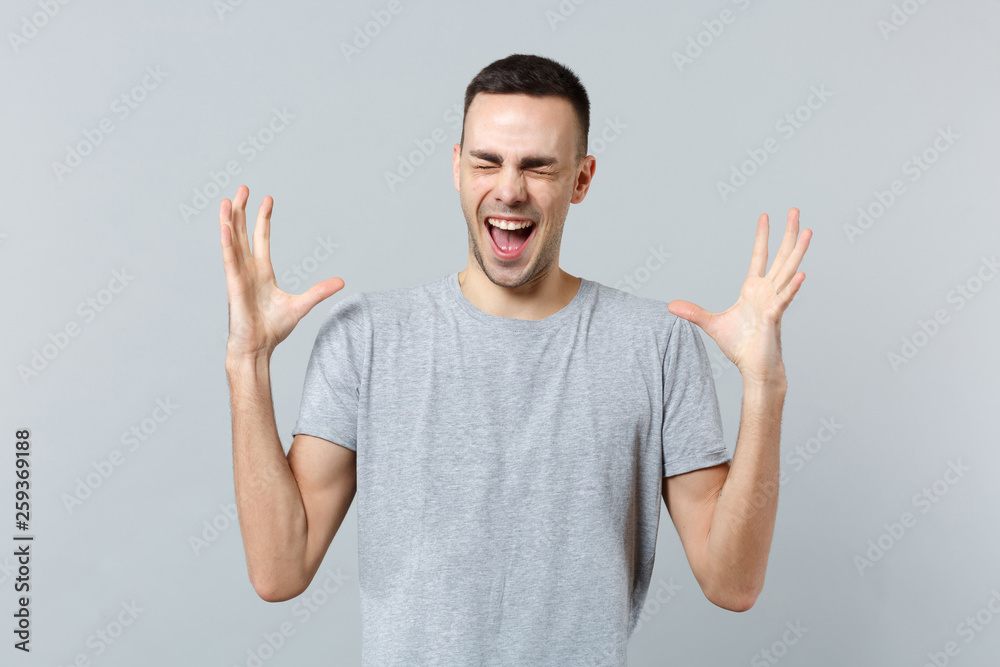 Portrait of crazy screaming young man in casual clothes keeping eyes closed, spreading hands isolated on grey wall background in studio. People sincere emotions, lifestyle concept. Mock up copy space. Portrait of crazy screaming young man in casual clothes keeping eyes closed, spreading hands isolated on grey wall background in studio. People sincere emotions, lifestyle concept. Mock up copy space.