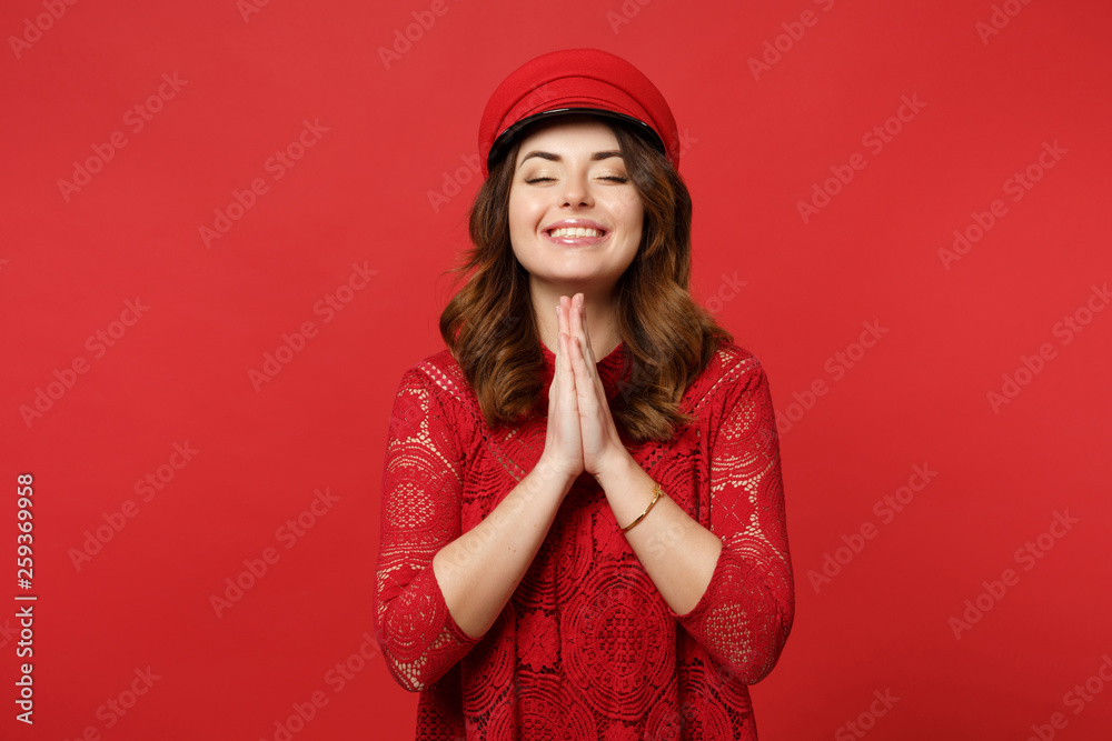 Portrait of smiling young woman in lace dress, cap keeping eyes closed, holding hands folded praying isolated on bright red background. People sincere emotions, lifestyle concept. Mock up copy space.
