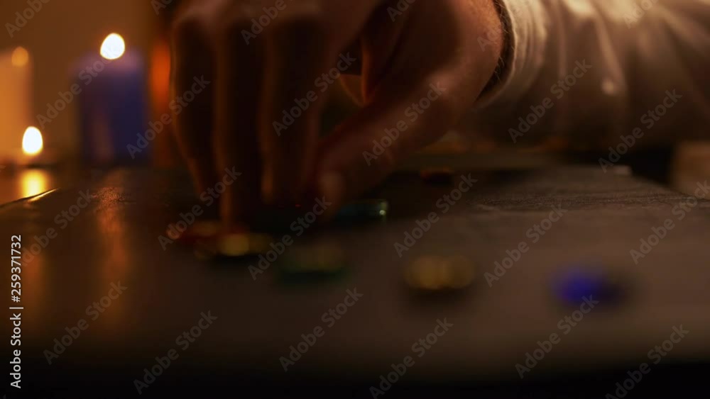 Close-up of a male hand in white, lying on a table, on black leather cardboard. A man lays out multicolored stones in a semicircle. In the background are burning candles. Side view. Close-up of a male hand in white, lying on a table, on black leather cardboard. A man lays out multicolored stones in a semicircle. In the background are burning candles. Side view.