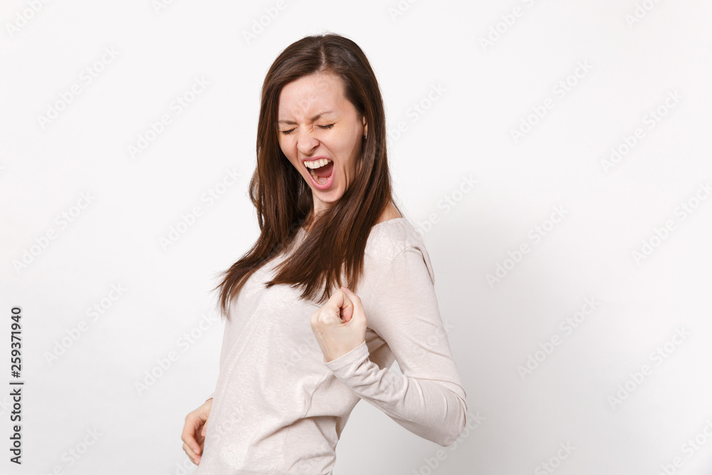 Portrait of screaming young woman in light clothes keeping eyes closed clenching fist like winner isolated on white background in studio. People sincere emotions lifestyle concept. Mock up copy space. Portrait of screaming young woman in light clothes keeping eyes closed clenching fist like winner isolated on white background in studio. People sincere emotions lifestyle concept. Mock up copy space.