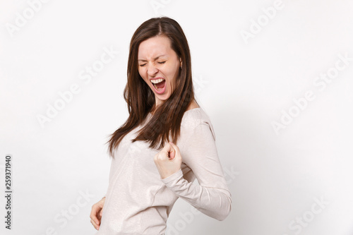 Portrait of screaming young woman in light clothes keeping eyes closed clenching fist like winner isolated on white background in studio. People sincere emotions lifestyle concept. Mock up copy space.