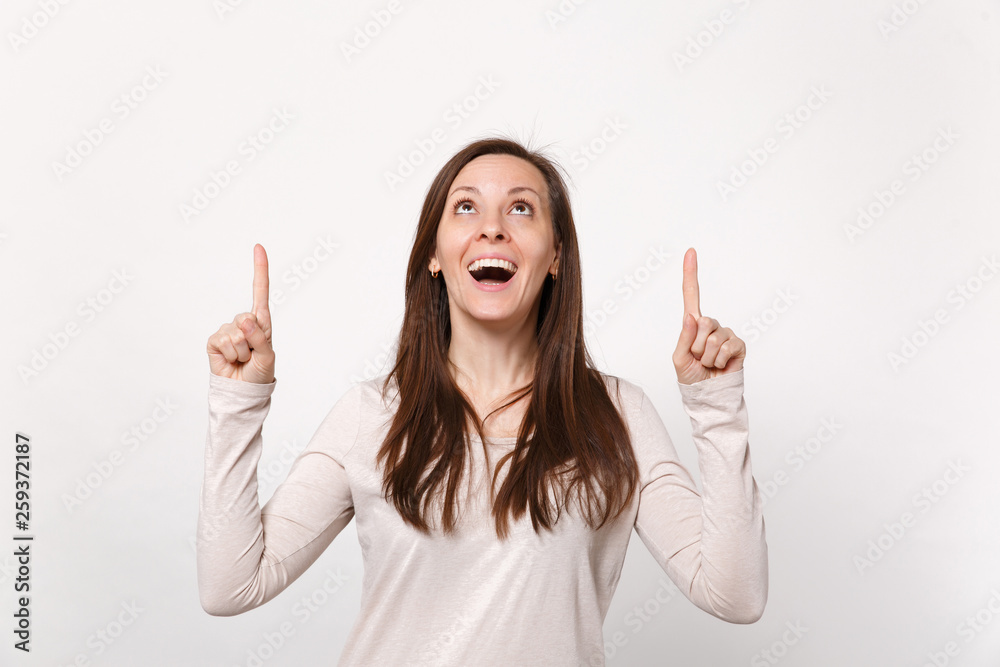 Portrait of cheerful excited young woman in light clothes looking pointing index fingers up isolated on white wall background in studio. People sincere emotions, lifestyle concept. Mock up copy space. Portrait of cheerful excited young woman in light clothes looking pointing index fingers up isolated on white wall background in studio. People sincere emotions, lifestyle concept. Mock up copy space.