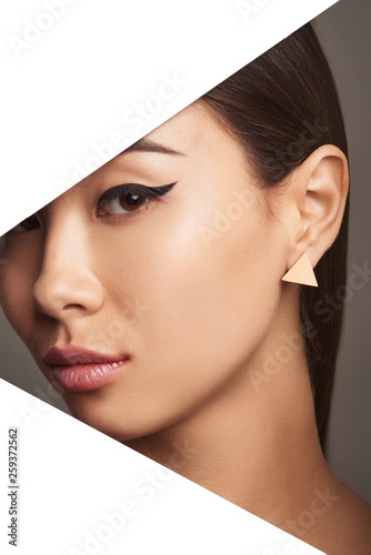 Cropped side half-turn geometric portrait of lady with plush lips and black flicks. The woman is wearing small beige triangle-shaped earring, looking at the camera behind triangle-shaped foreground.