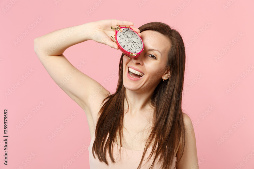 Smiling young woman covering eye with half of fresh ripe pitahaya, dragon fruit isolated on pink pastel wall background in studio. People vivid lifestyle relax vacation concept. Mock up copy space. Smiling young woman covering eye with half of fresh ripe pitahaya, dragon fruit isolated on pink pastel wall background in studio. People vivid lifestyle relax vacation concept. Mock up copy space.