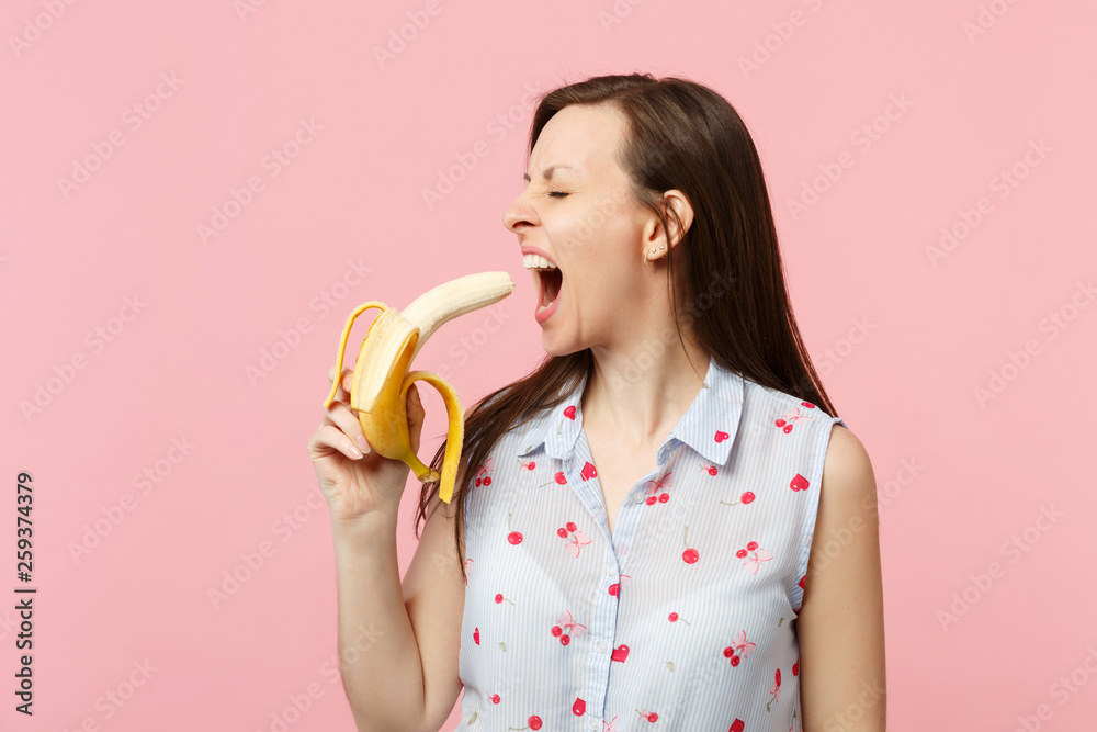 Crazy young woman in summer clothes keeping eyes closed biting fresh ripe banana fruit isolated on pink pastel background in studio. People vivid lifestyle, relax vacation concept. Mock up copy space. Crazy young woman in summer clothes keeping eyes closed biting fresh ripe banana fruit isolated on pink pastel background in studio. People vivid lifestyle, relax vacation concept. Mock up copy space.