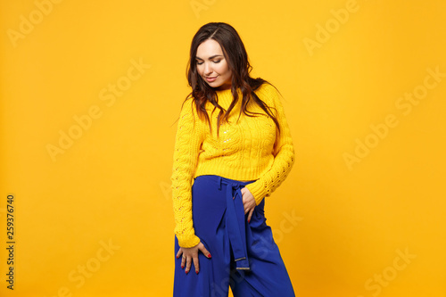 Portrait of tender young woman in sweater, blue trousers standing and looking down isolated on yellow orange wall background in studio. People sincere emotions, lifestyle concept. Mock up copy space.