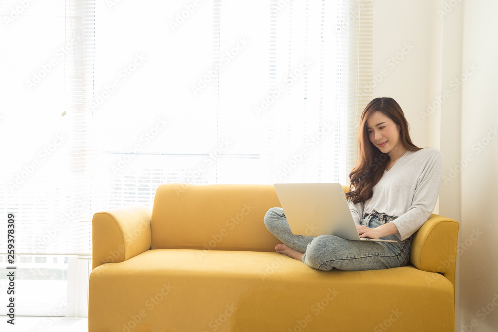 Young Asian woman sitting with laptop on yellow couch in home office. Concept of remote workplace and working at home Young Asian woman sitting with laptop on yellow couch in home office. Concept of remote workplace and working at home