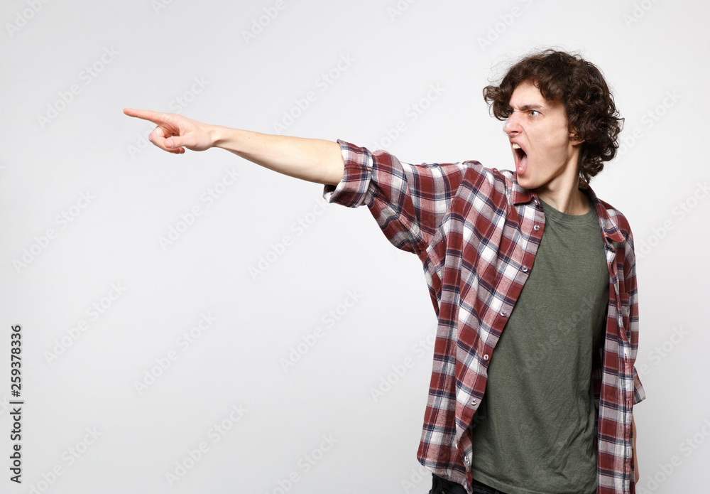 Portrait of angry screaming young man in casual clothes swearing, looking pointing index finger aside isolated on white wall background. People sincere emotions, lifestyle concept. Mock up copy space.