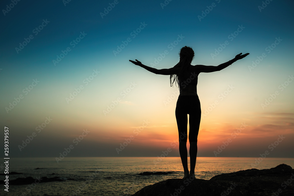 Silhouette of a flexible yoga woman stands on the shore of the ocean at evening.
