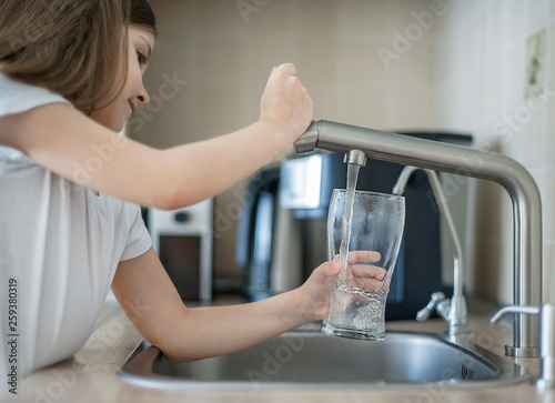 Portrait of a little caucasian girl gaining a glass of tap clean water. Kitchen faucet. Cute kid pouring fresh water from filter tap. Indoors. Healthy life concept