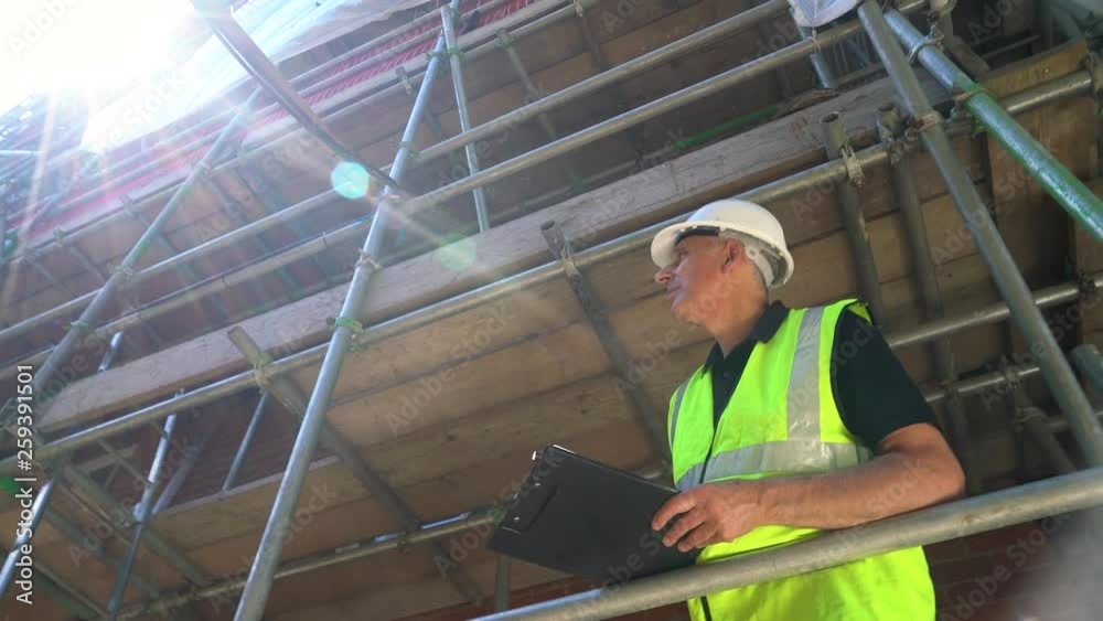 Moving camera clip of male builder foreman, surveyor, construction worker, surveyor, or architect working on construction building site standing on scaffolding writing on clipboard 