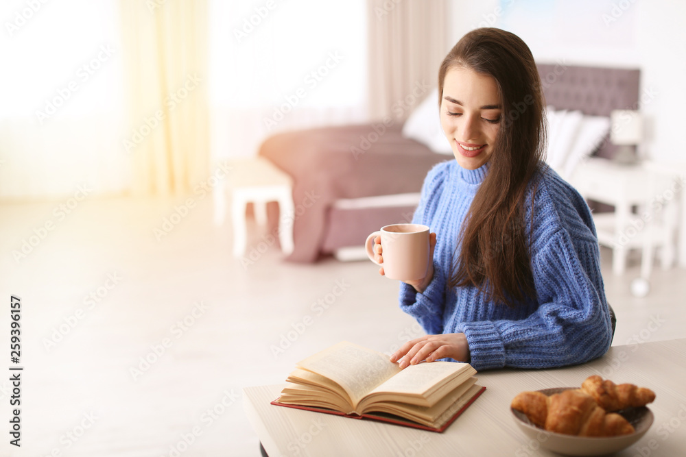 Young woman drinking coffee and reading book at table indoors, space for text Young woman drinking coffee and reading book at table indoors, space for text