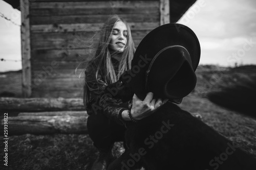 Happy young woman plaing with her black dog in fron of old wooden house. Girl tries a hat to her dog. Black and white photo photo