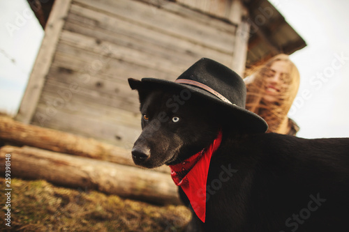 Happy young woman plaing with her black dog in fron of old wooden house. Girl tries a hat to her dog photo