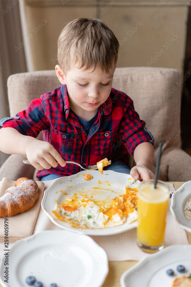 Funny and happy boy having Breakfast. Light brunch near window in a cafe. Croissants, omelet, orange juice and many different dishes on the table in the cafe. Funny and happy boy having Breakfast. Light brunch near window in a cafe. Croissants, omelet, orange juice and many different dishes on the table in the cafe.