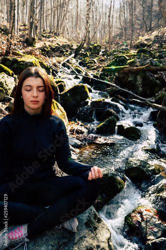 young sporty european girl making Yoga Pose lotus Sitting in a rock on a river Meditating in the forest with sunlight