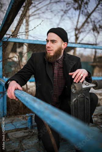 Bearded Skater waring Black Coat and Black Hat Holds His Skateboard and Sits Relaxing
