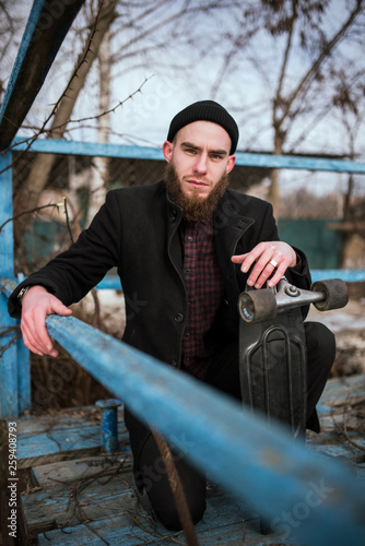 Bearded Skater waring Black Coat and Black Hat Holds His Skateboard and Sits Relaxing © Andrii