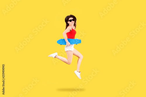 Full length portrait of a girl in sunglasses with skateboard jumping on yellow background, youth summer concept