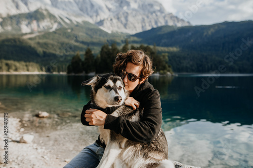 Mensch und Hund umarmen sich an einem schönen See mit Bergen im Hintergrund - Freundschaft