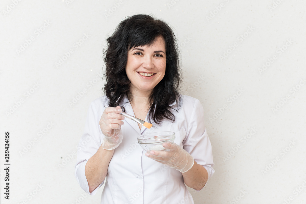 advertising campaign for cosmetology. A girl in a white medical gown mixes a mask on a white background and applies it to her face. Smile and emotions advertising campaign for cosmetology. A girl in a white medical gown mixes a mask on a white background and applies it to her face. Smile and emotions