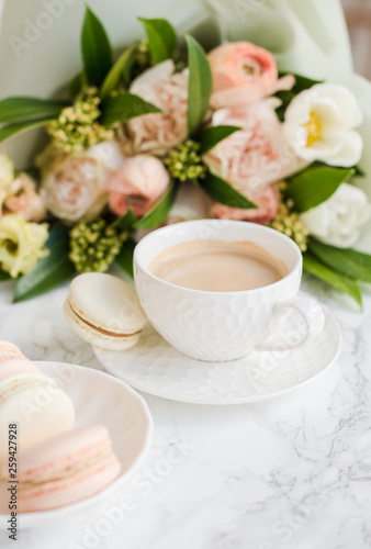Elegant sweet dessert macarons, cup of coffee and pastel colored beige flowers bouquet on white marble