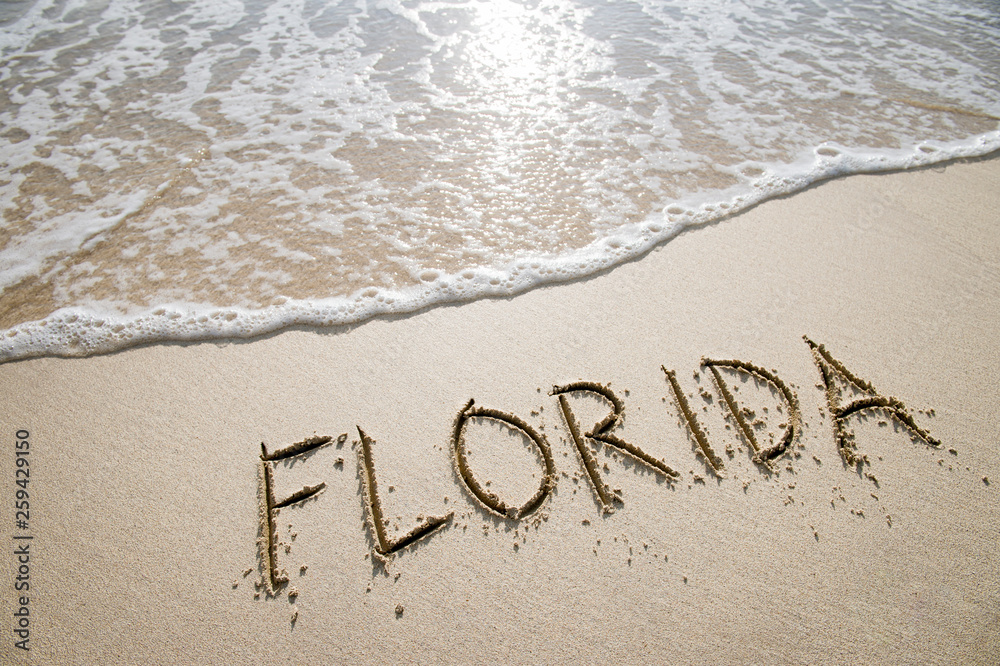 Fototapeta premium Simple Florida American holiday getaway message written in smooth sand with an incoming wave on a tropical beach