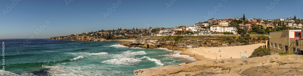 Sydney, Australia - February 11, 2019: Panorama shot of sandy Tamarama beach in front and Bronte Beach in distance. Rochy shorelines and plenty of housing. Blue sky and sea.