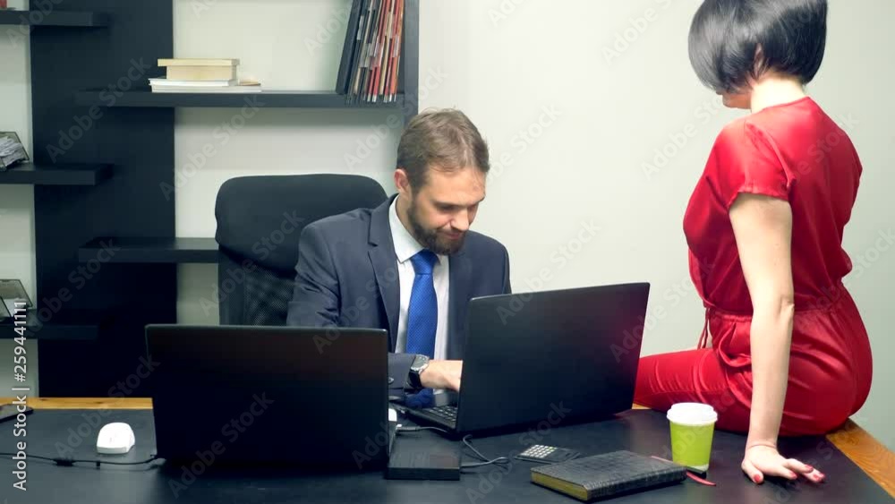 Office flirt. An attractive woman in a red jumpsuit is sitting on the desk of her colleague and sensually touches his glass of coffee.
