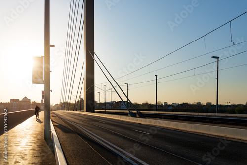 Silhouette view against sunlight of cyclist ride bicycle on the pedestrian pathway on suspension bridge with railway at the centre and background of cityscape and gradient blue and orange sky.