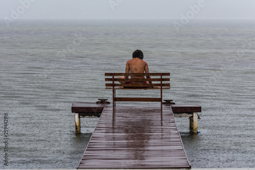 A man sits on a bench in front of a lake during a rainy day.