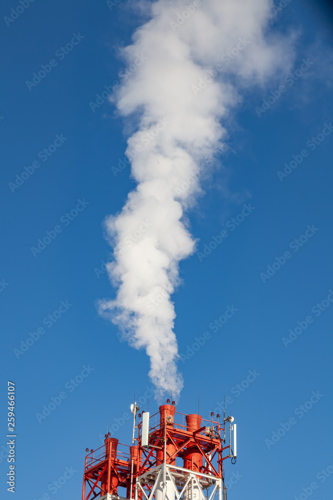 White harmful smoke coming out of the red-and-white pipes with mobile communication antennas at a factory in the city center against the background of a clear blue sky. Environmental problems.