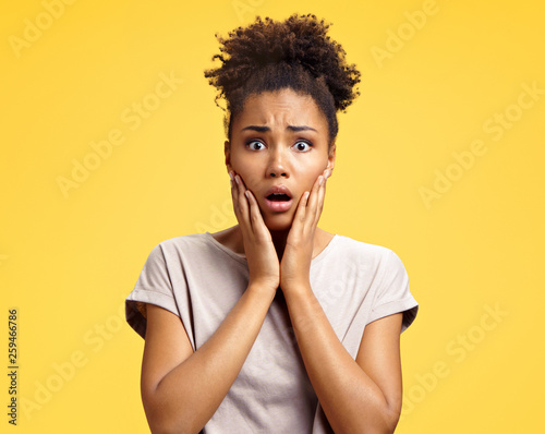 Astonished girl with big eyes, opens mouth widely, keeps hands on cheeks. Photo of african american girl wears casual outfit on yellow background. Emotions and Omg concept