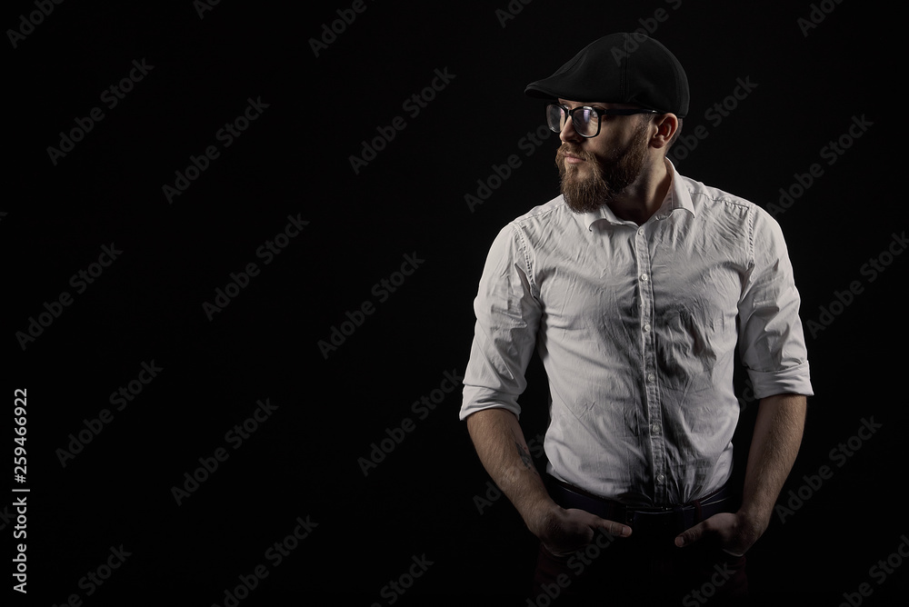 Portrait of a young man in glasses with a beard and a mustache in a black cap and a white shirt, A dark studio background