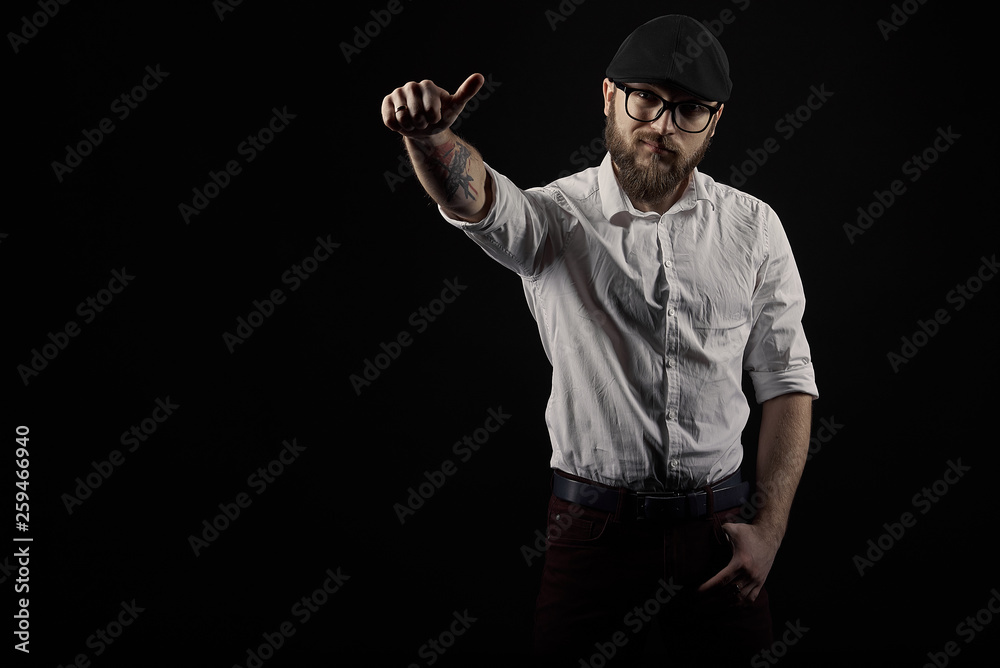 young handsome man in glasses with a beard and a mustache in a black cap and a white shirt shows a variety of hand gestures, a dark studio background