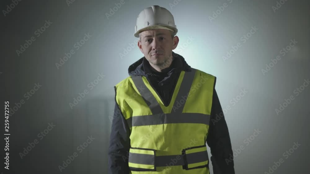 Portrait of the worker in the builders uniform and helmet on a black background in the spotlight at the workplace. Portrait of the worker in the builders uniform and helmet on a black background in the spotlight at the workplace.