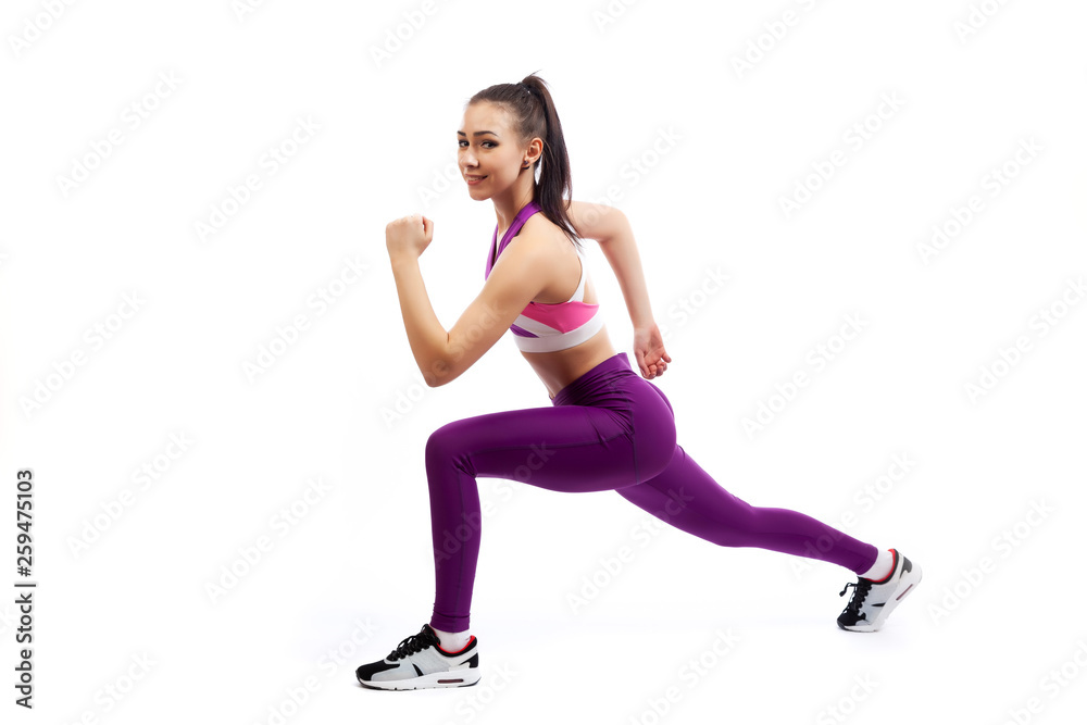A young  woman coach in a sporty short top and gym leggings makes lunges  by the feet forward, hands are held out to the side   on a  white isolated background in studio