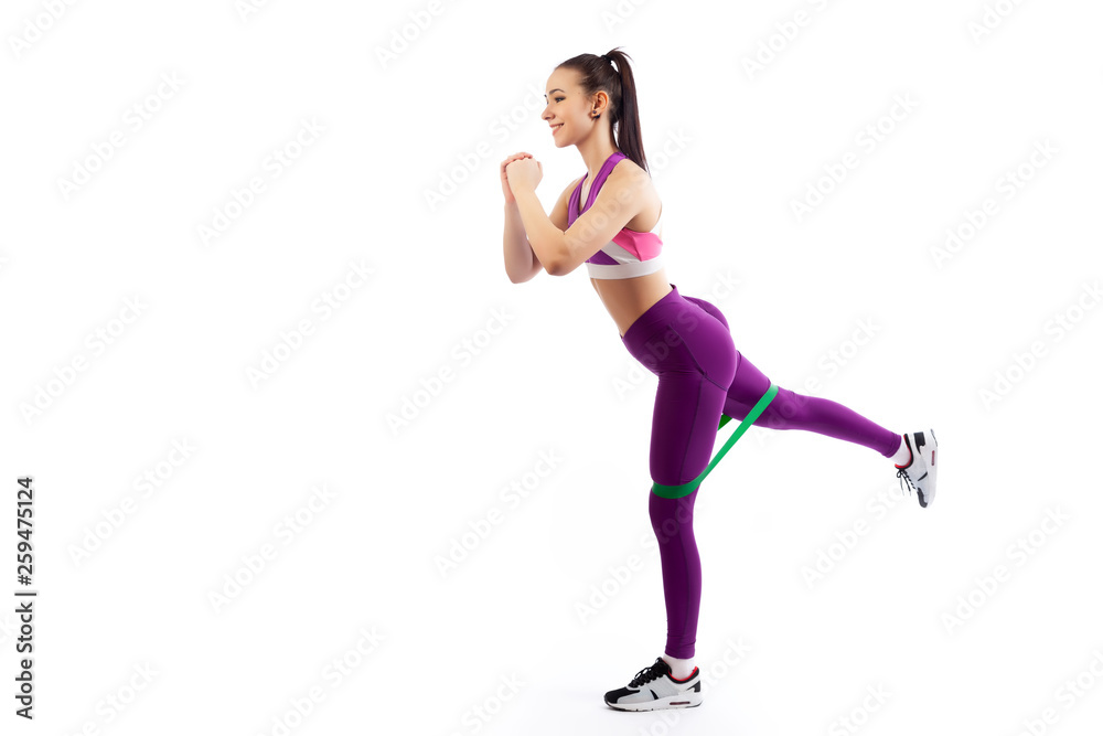 A young  woman coach in a sporty  short top and gym shows the correct makes lunges with sport fitness rubber bands on a  white isolated background in studio