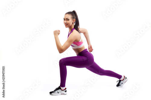 A young woman coach in a sporty short top and gym leggings makes lunges by the feet forward, hands are held out to the side on a white isolated background in studio