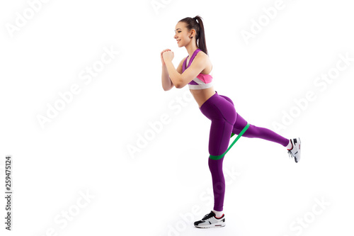 A young woman coach in a sporty short top and gym shows the correct makes lunges with sport fitness rubber bands on a white isolated background in studio